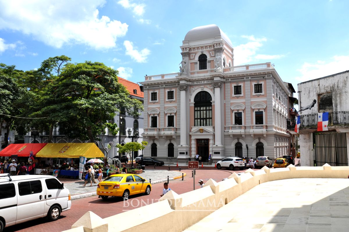 Ein historisches weißes Gebäude steht auf einem belebten Platz in Panama, umgeben von Marktständen, Taxis, Menschen und Bäumen.