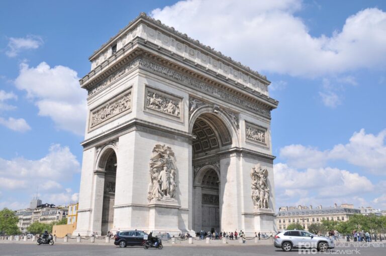 Der Arc de Triomphe in Paris steht unter blauem Himmel, umgeben von Autos und Motorrädern auf den belebten Straßen.