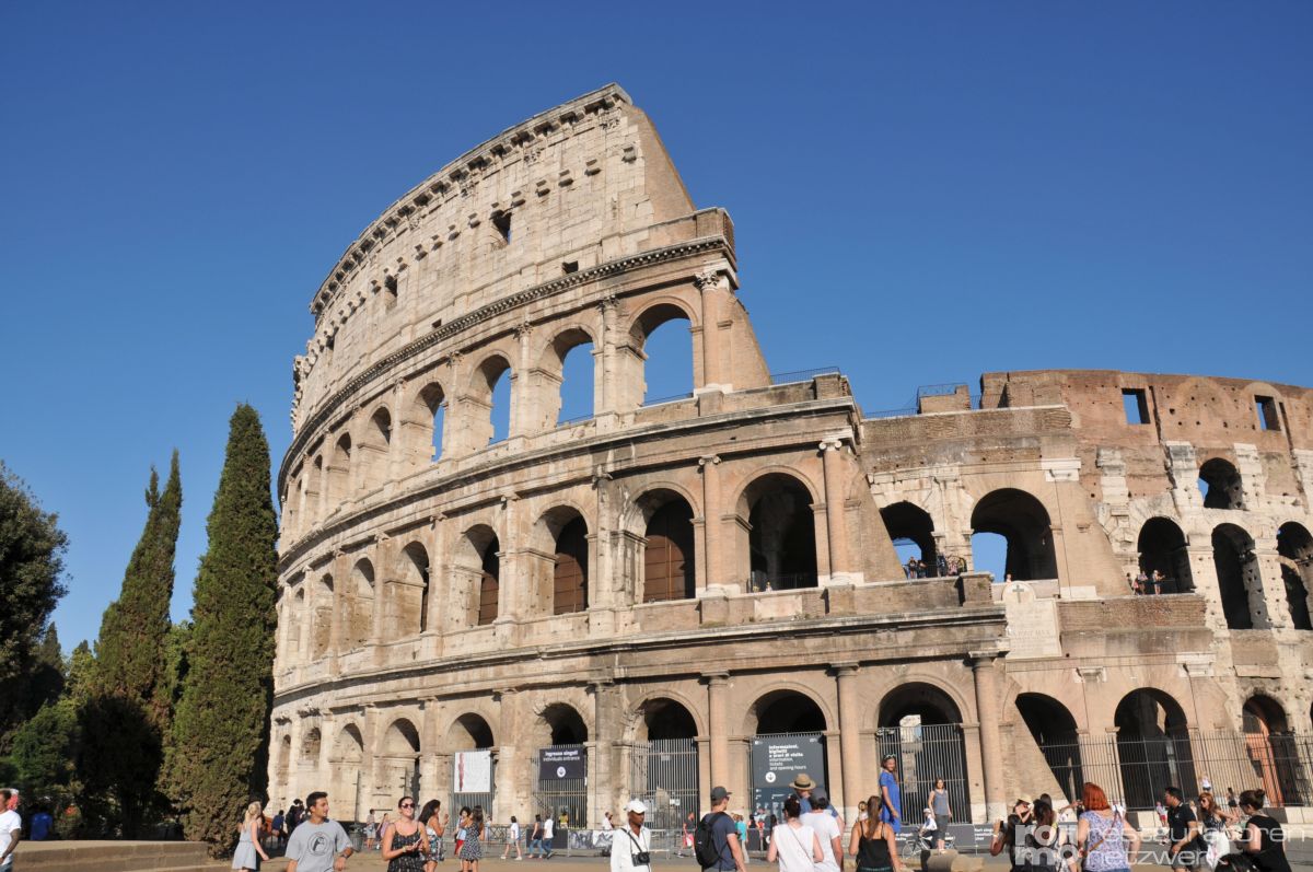 Das Kolosseum in Rom steht unter blauem Himmel, umgeben von Touristen und grünen Zypressen.