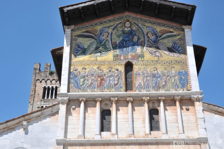 Die Fassade einer Kirche in Lucca zeigt ein Mosaik mit religiösen Figuren, Säulen, Rundbogenfenstern und links einen Glockenturm.