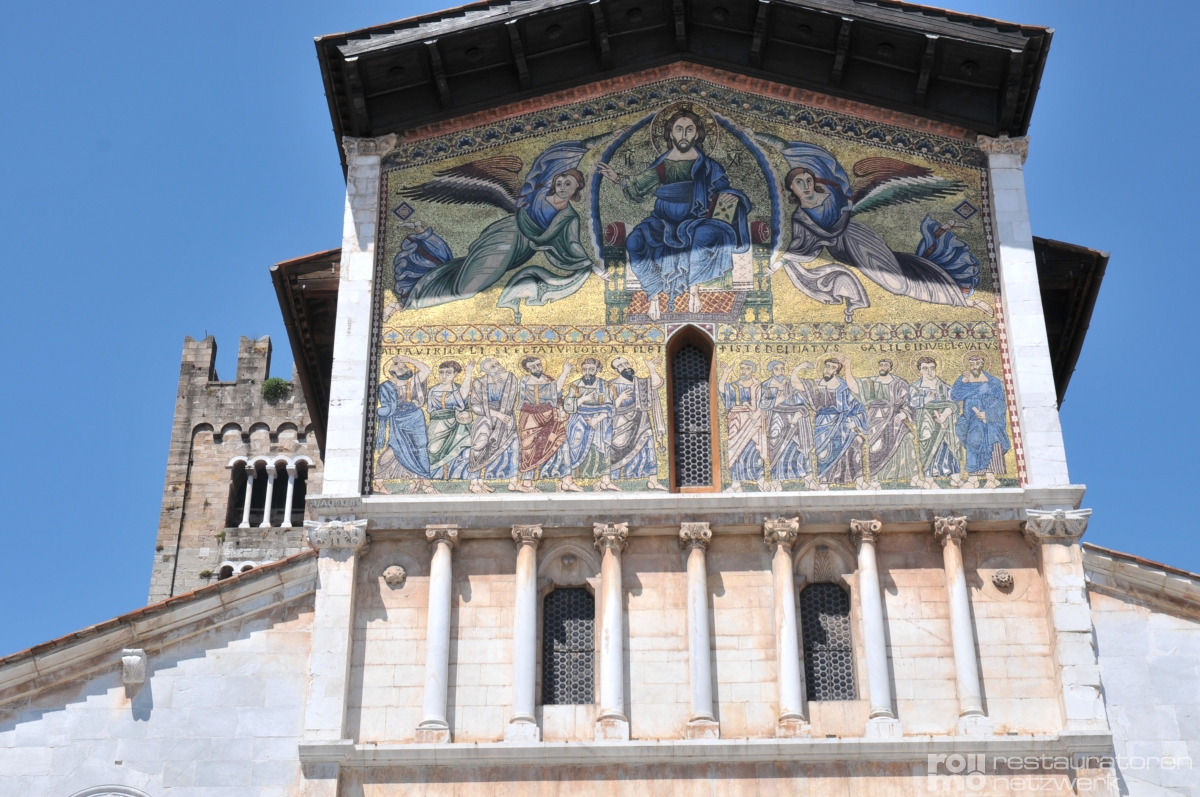 Die Fassade einer Kirche in Lucca zeigt ein Mosaik mit religiösen Figuren, Säulen, Rundbogenfenstern und links einen Glockenturm.