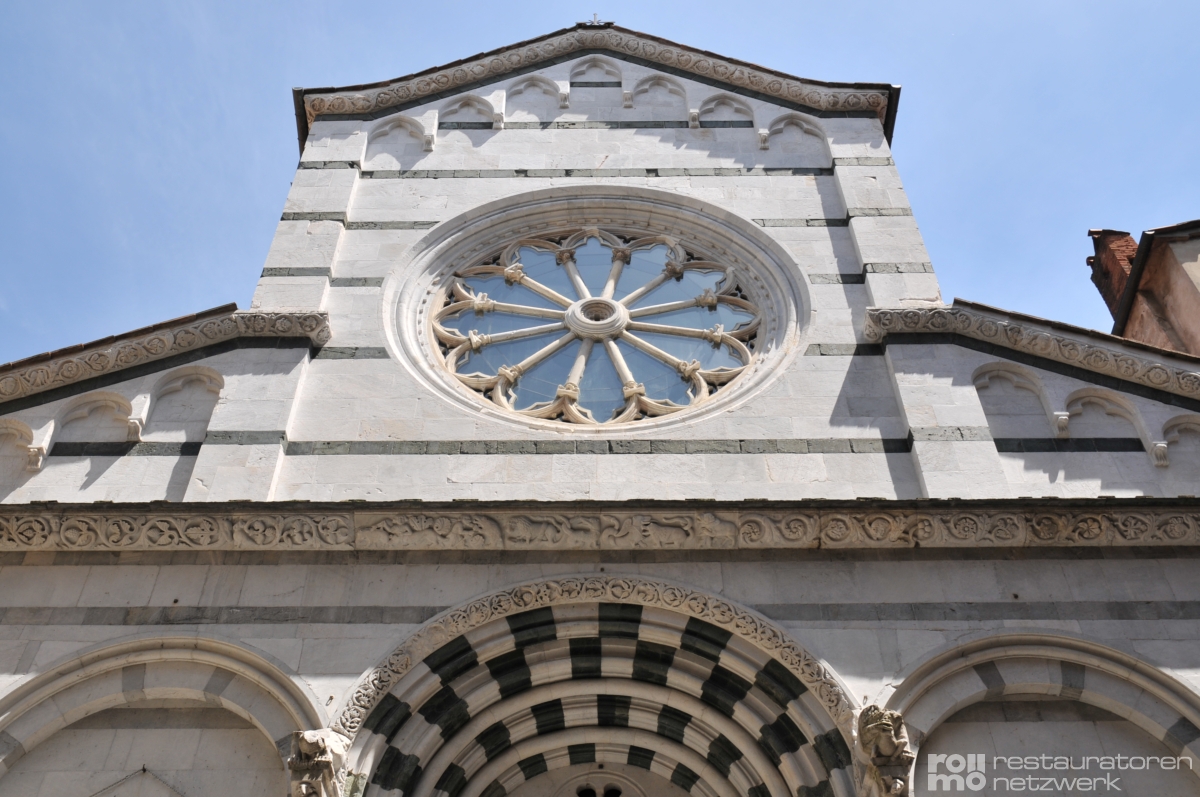 Die Fassade zeigt eine Steinkirche mit Streifen, großem Rundfenster und verzierten Bögen über dem Eingang.