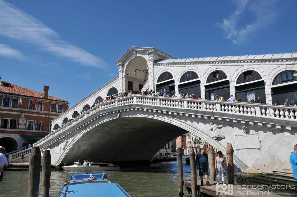 Das Bild zeigt die Rialtobrücke in Venedig über dem Canal Grande, mit Fußgängern und einem blauen Boot im Wasser.