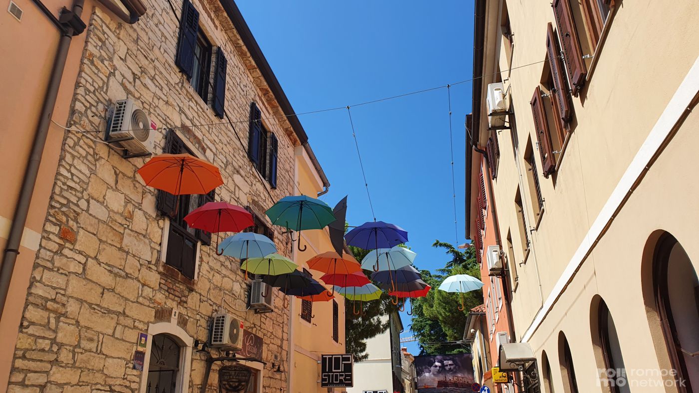 Bunte Regenschirme bilden ein Dach über einer engen Gasse mit Stein- und Stuckgebäuden unter blauem Himmel.
