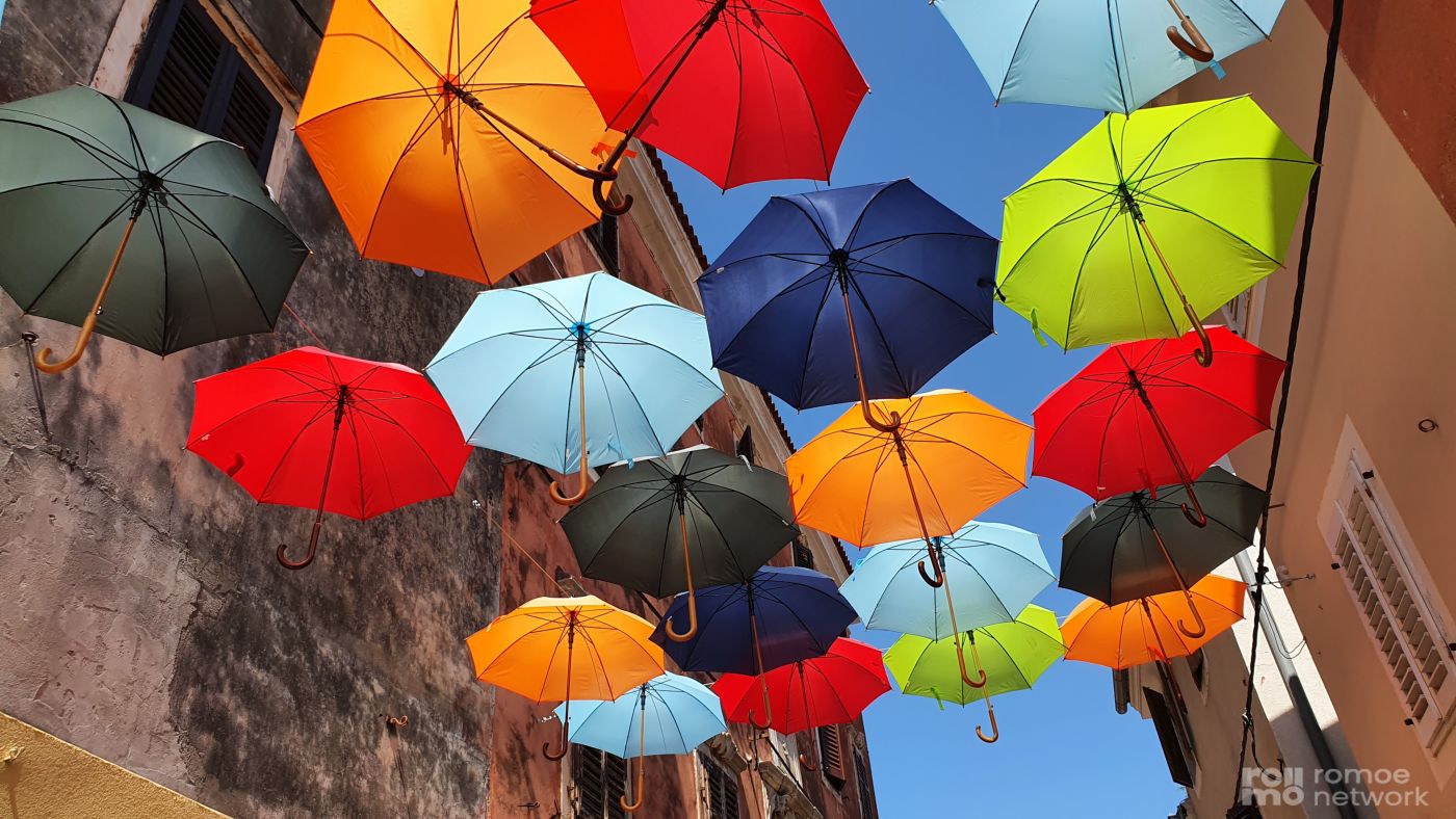 Bunte Regenschirme hängen über einer schmalen Straße und bilden ein lebendiges Dach vor blauem Himmel.
