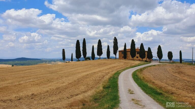 Ein Feldweg führt durch ein goldenes Feld zu einem Bauernhaus in der Toskana mit Zypressen unter teils bewölktem Himmel.
