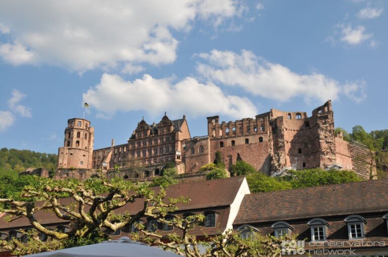 Das Heidelberger Schloss, teilweise ruinös, thront auf einem grünen Hügel über roten Dächern unter bewölktem Himmel.