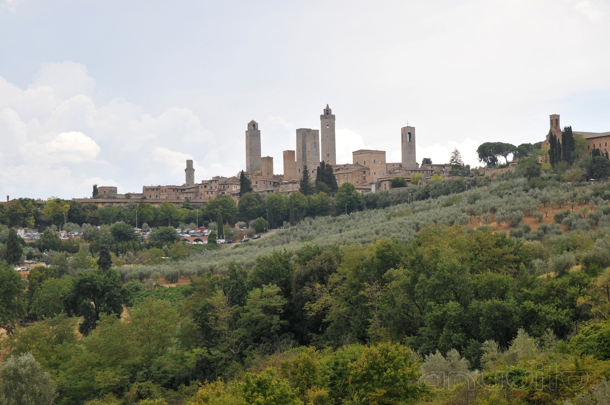 San Gimignano, Italien: mittelalterliche Türme über steinernen Gebäuden, mit grünen Bäumen und sanften Hügeln unter einem teilweise bewölkten Himmel.