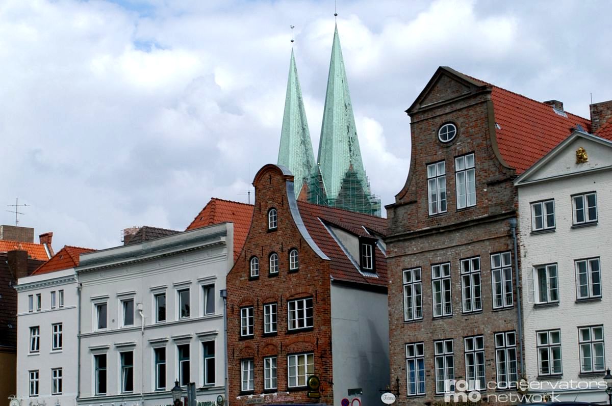 Historische europäische Gebäude mit roten und weißen Fassaden stehen vor zwei hohen grünen Kirchtürmen unter einem teilweise bewölkten Himmel.