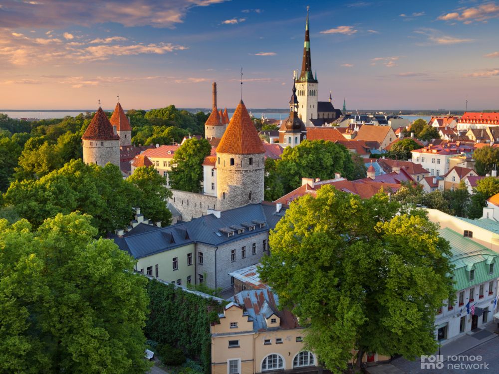 Tallinn mit roten Turmdächern, historischen Gebäuden, grünen Bäumen und Kirchturm bei teils bewölktem Himmel im Sonnenuntergang.