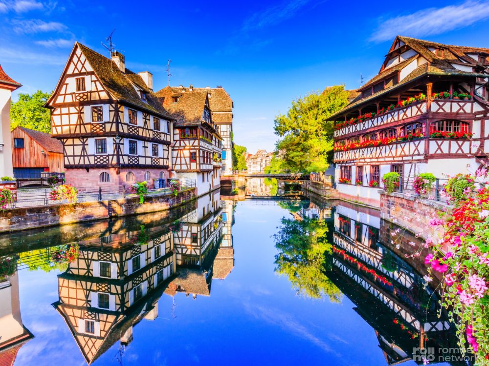 Fachwerkhäuser, die sich in einem ruhigen Kanal, blauem Himmel, Grünpflanzen und Blumenkästen in Straßburg, Frankreich, spiegeln.