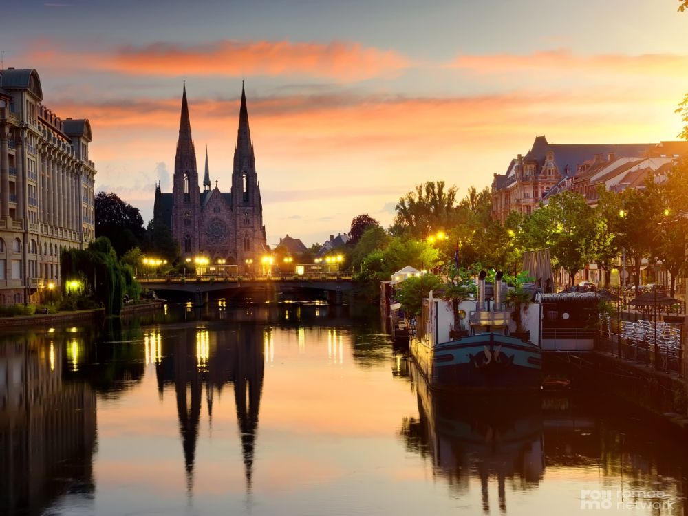 Sonnenuntergang am Fluss mit Kathedrale (zwei Türme), europäischen Gebäuden und einem Boot im Wasser, das den Himmel bespiegelt.