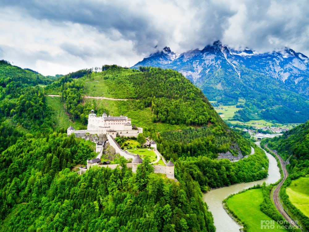 Luftaufnahme eines weißen Schlosses inmitten von grünen Wäldern und Hügeln, mit einem Fluss im Hintergrund und schneebedeckten Bergen.