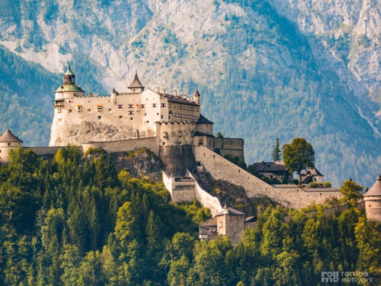 Mittelalterliche Burg mit mehreren Türmen auf bewaldetem Hügel, umgeben von Mauern, vor hohen Bergen und klarem Himmel.