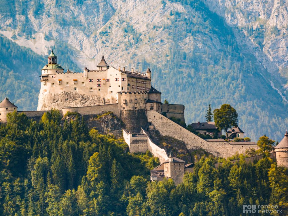 Mittelalterliche Burg mit mehreren Türmen auf bewaldetem Hügel, umgeben von Mauern, vor hohen Bergen und klarem Himmel.