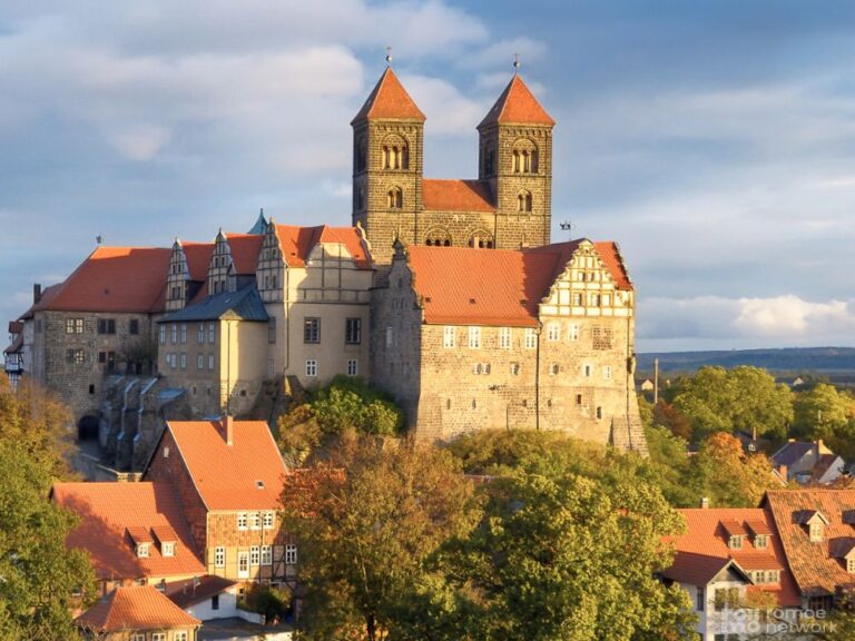 Quedlinburg - Historische Steinfestung mit zwei Türmen und roten Dächern über einer Stadt, umgeben von Herbstbäumen unter teils bewölktem Himmel.