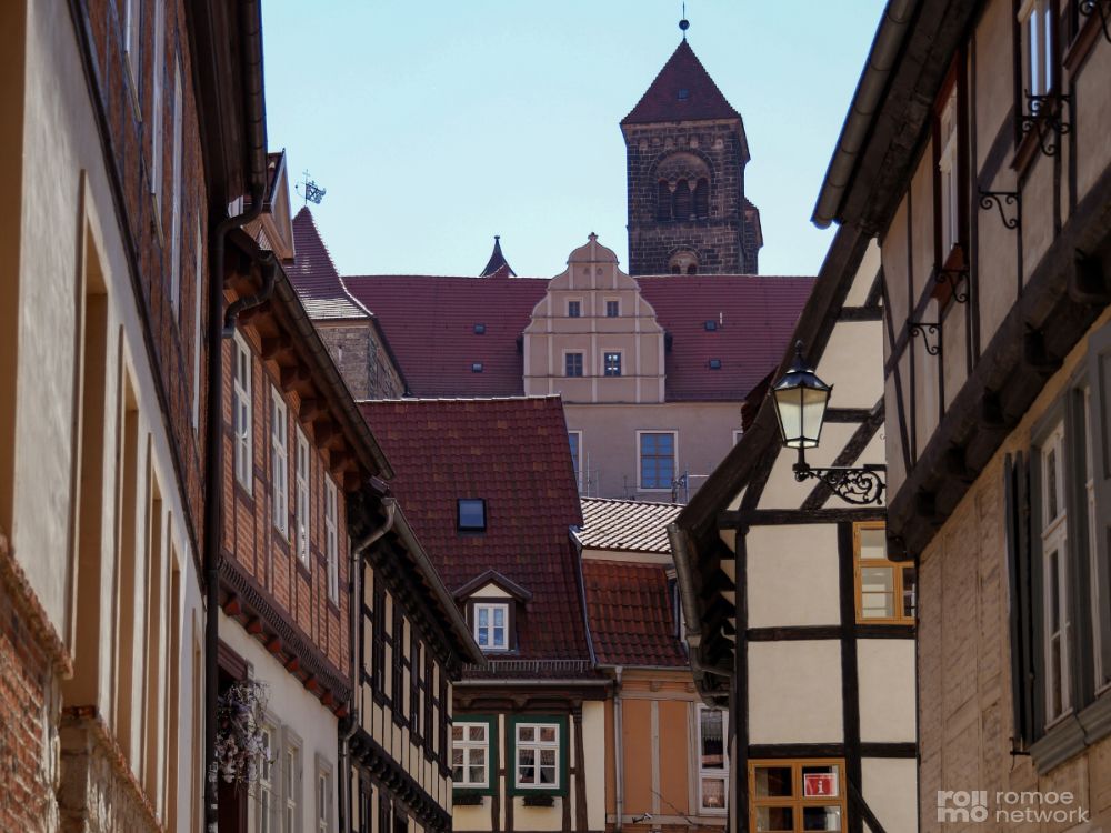 Quedlinburg - Schmale Straße mit historischen Fachwerkhäusern, die zu einem Kirchturm und einem verschnörkelten Gebäude unter freiem Himmel führt.