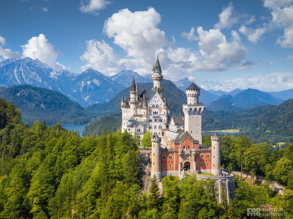 Schloss Neuschwanstein in Deutschland auf einem bewaldeten Hügel, mit Bergen, einem See und blauem Himmel im Hintergrund.
