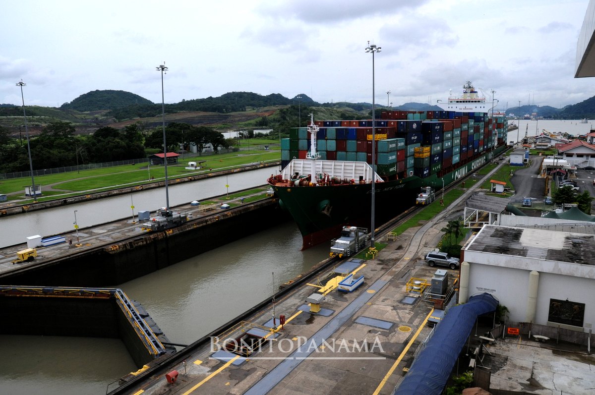Frachtschiff mit Containern in der Schleuse des Panamakanals, umgeben von grünen Hügeln, Wasser und Industriegebäuden unter Wolken.