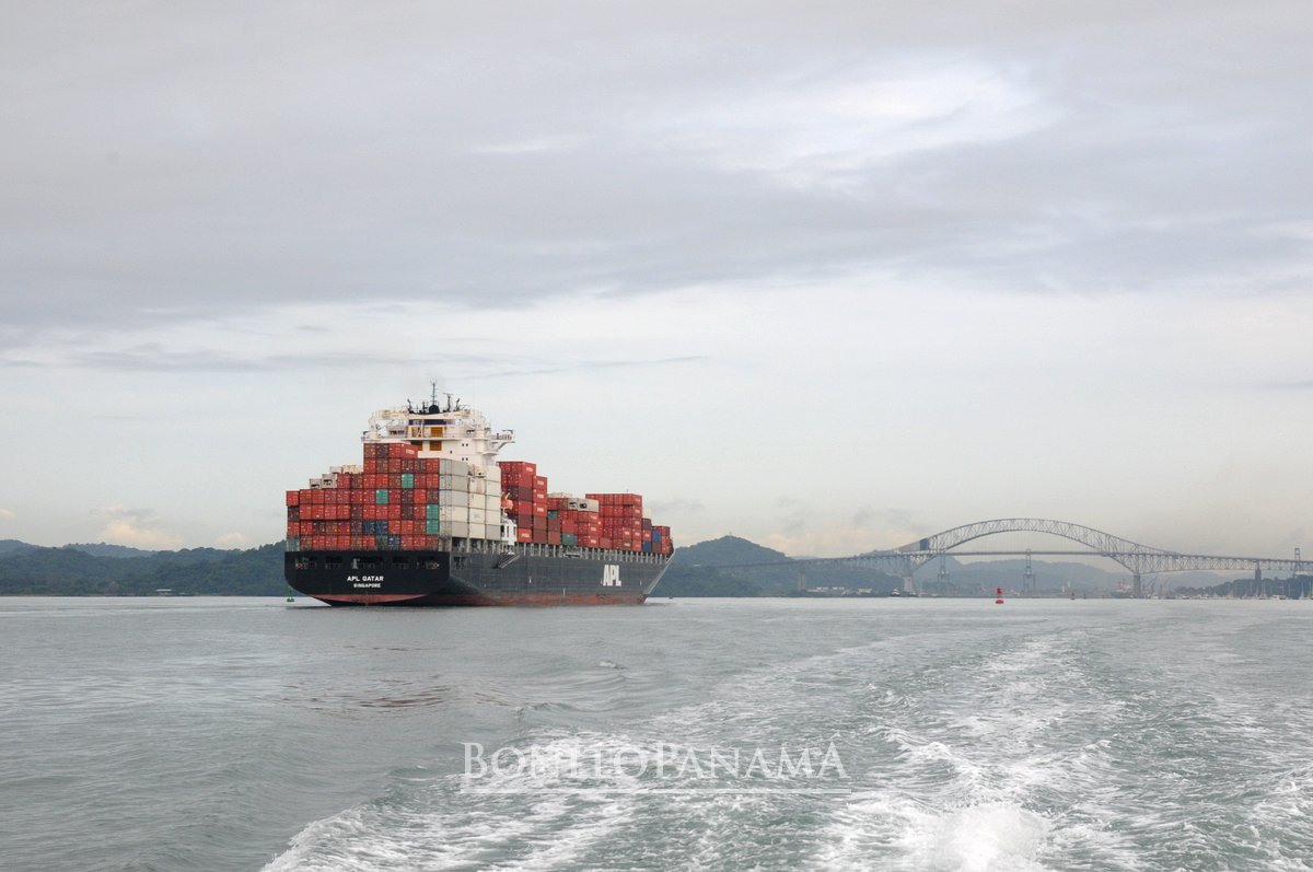 Großes Containerschiff auf Wasserweg, gestapelte Container an Bord, Brücke im Hintergrund, Land an beiden Seiten - BonitoPanama