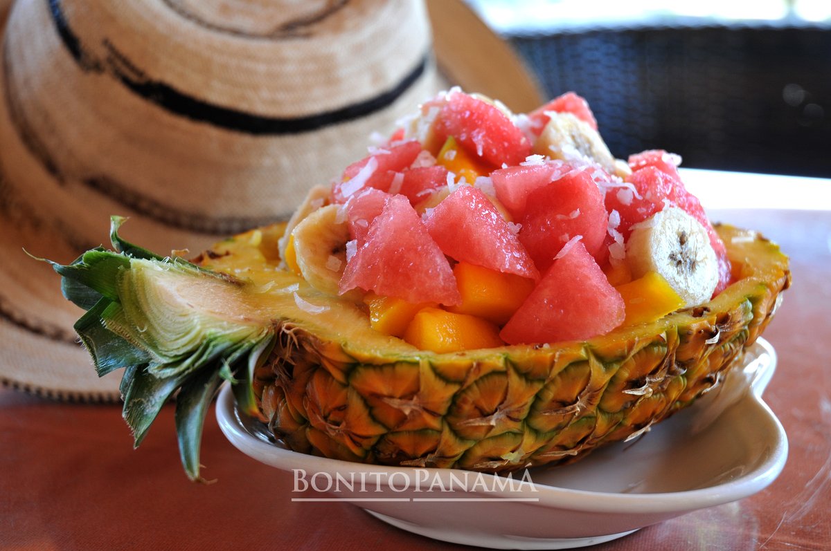 Halbe Ananas als Schale mit Obst und Kokos auf Teller, daneben eine geflochtene Panama-Hütte.