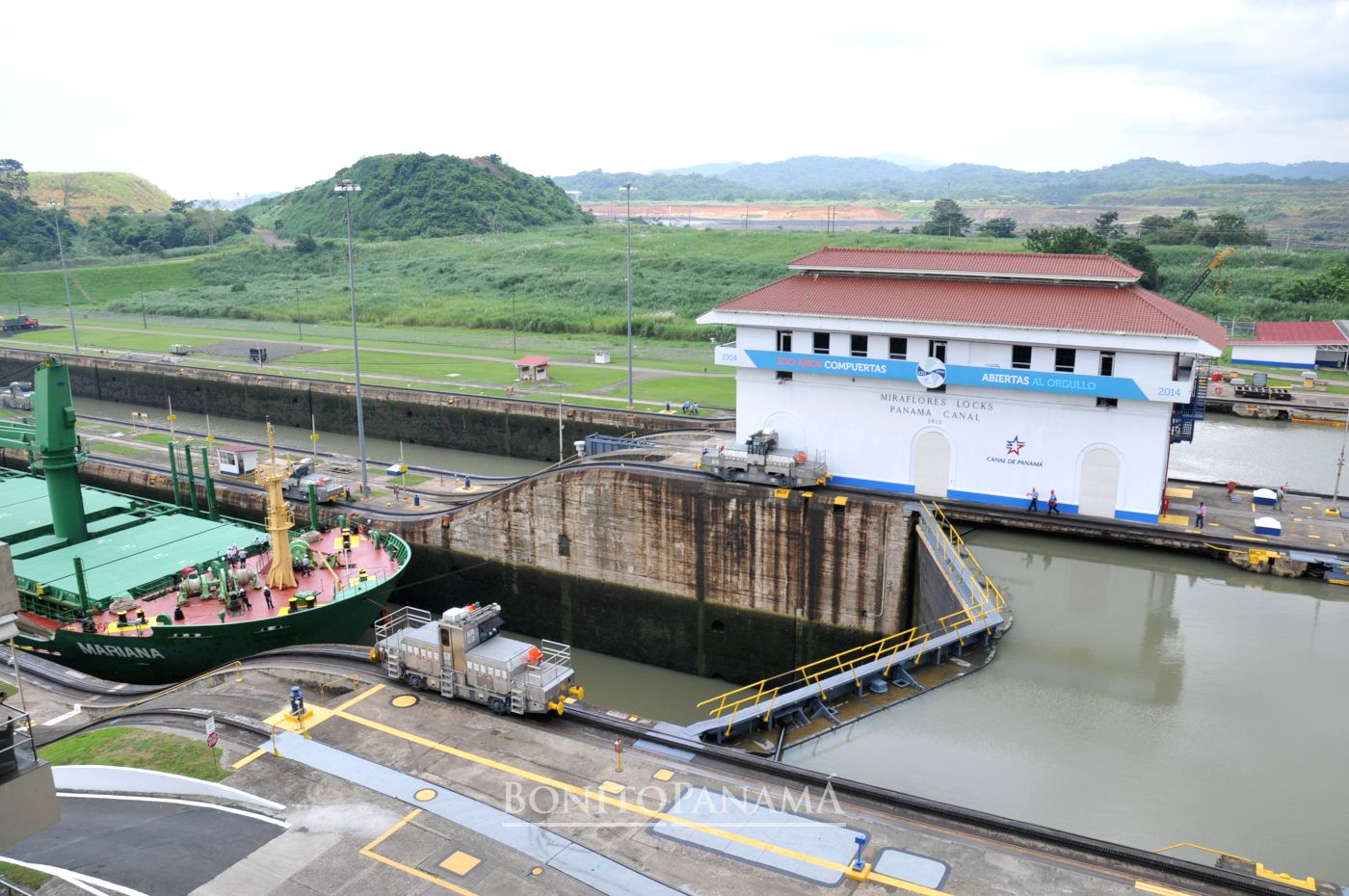 Frachtschiff in den Miraflores-Schleusen, Panamakanal; weißes Kontrollgebäude und grüne Hügel im Hintergrund.