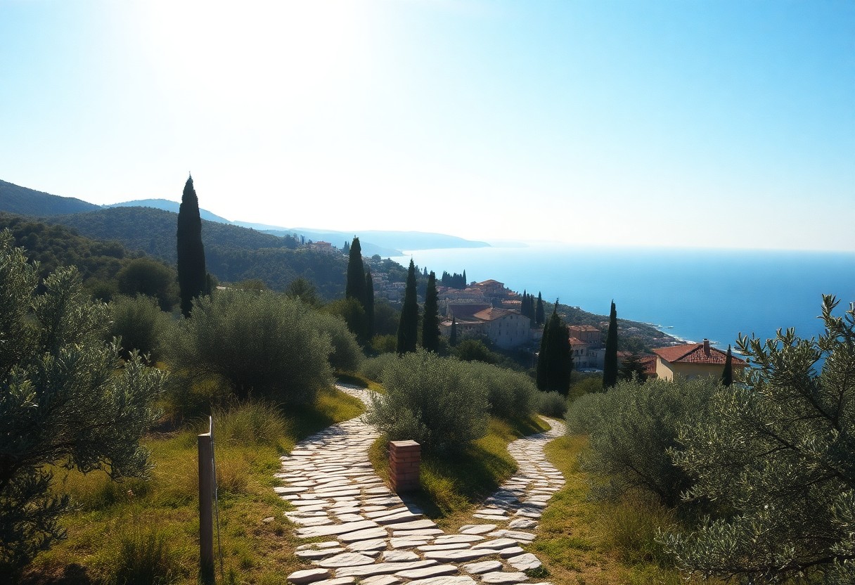 Steinweg mit Olivenbäumen und Zypressen führt zu terrakottabedeckten Häusern am Meer, Berge und blauer Himmel im Hintergrund.