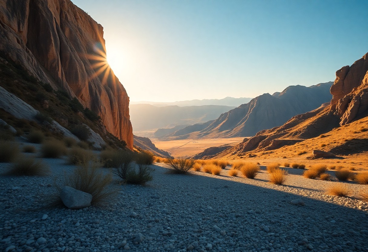 Sonnenschein über einer Schlucht mit Felsen, trockenem Gras im Vordergrund und Bergen unter klarem Himmel im Hintergrund.